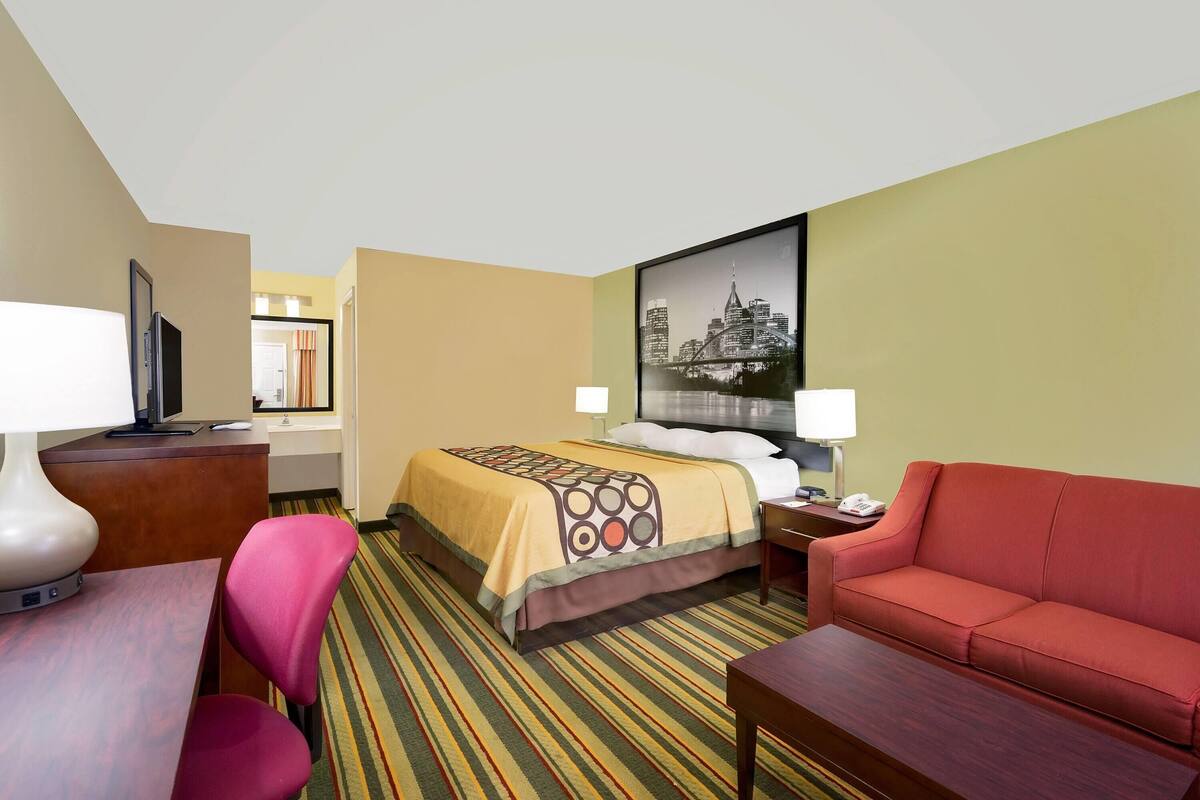 A hotel room with a bed, a desk, a red sofa, a TV, and a cityscape picture on the wall.