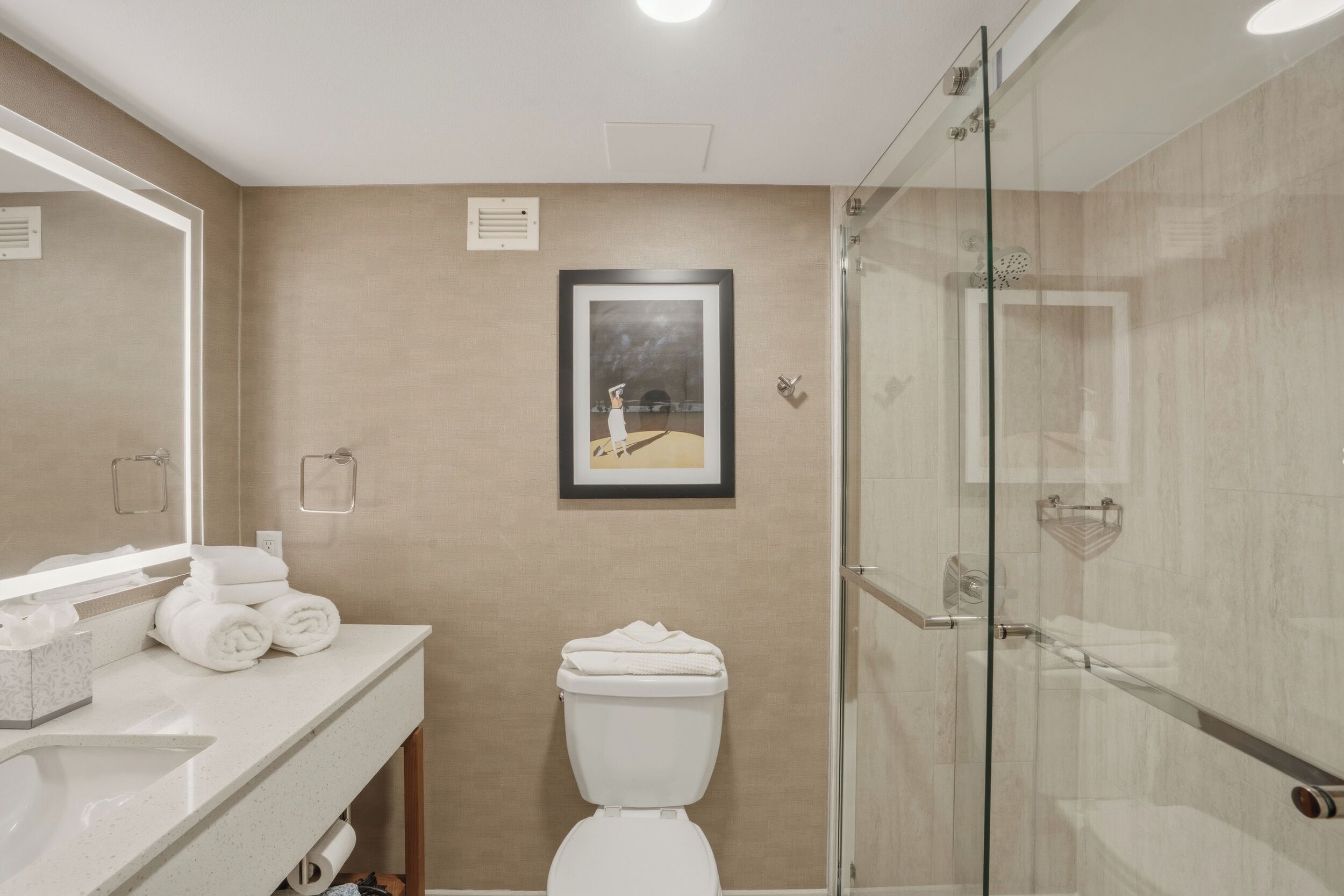 A bathroom with a glass shower enclosure, a toilet, a sink with a mirror, and a framed picture on the wall.