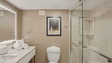 A bathroom with a glass shower enclosure, a toilet, a sink with a mirror, and a framed picture on the wall.