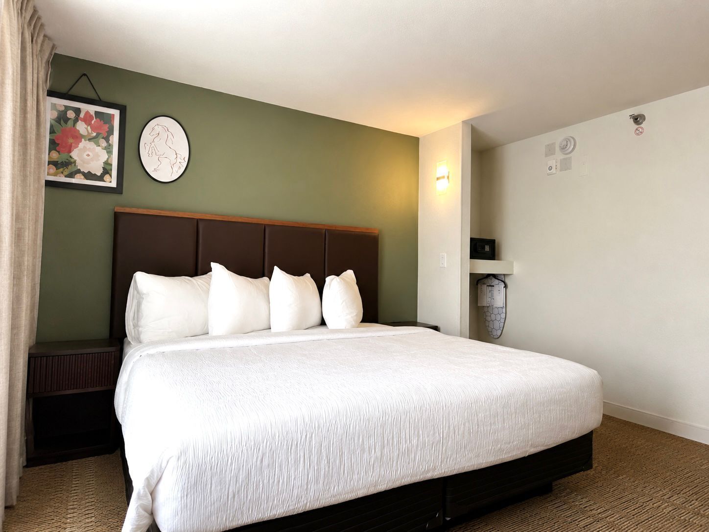 A hotel room with a large bed, a headboard, a wall clock, a framed floral artwork, and a wall-mounted light.