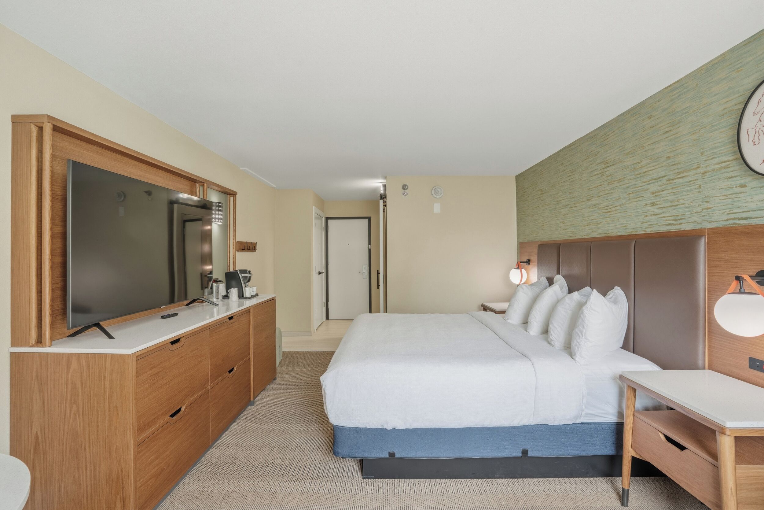 A hotel room with a large bed, a wooden dresser, a television, and a desk with a lamp.