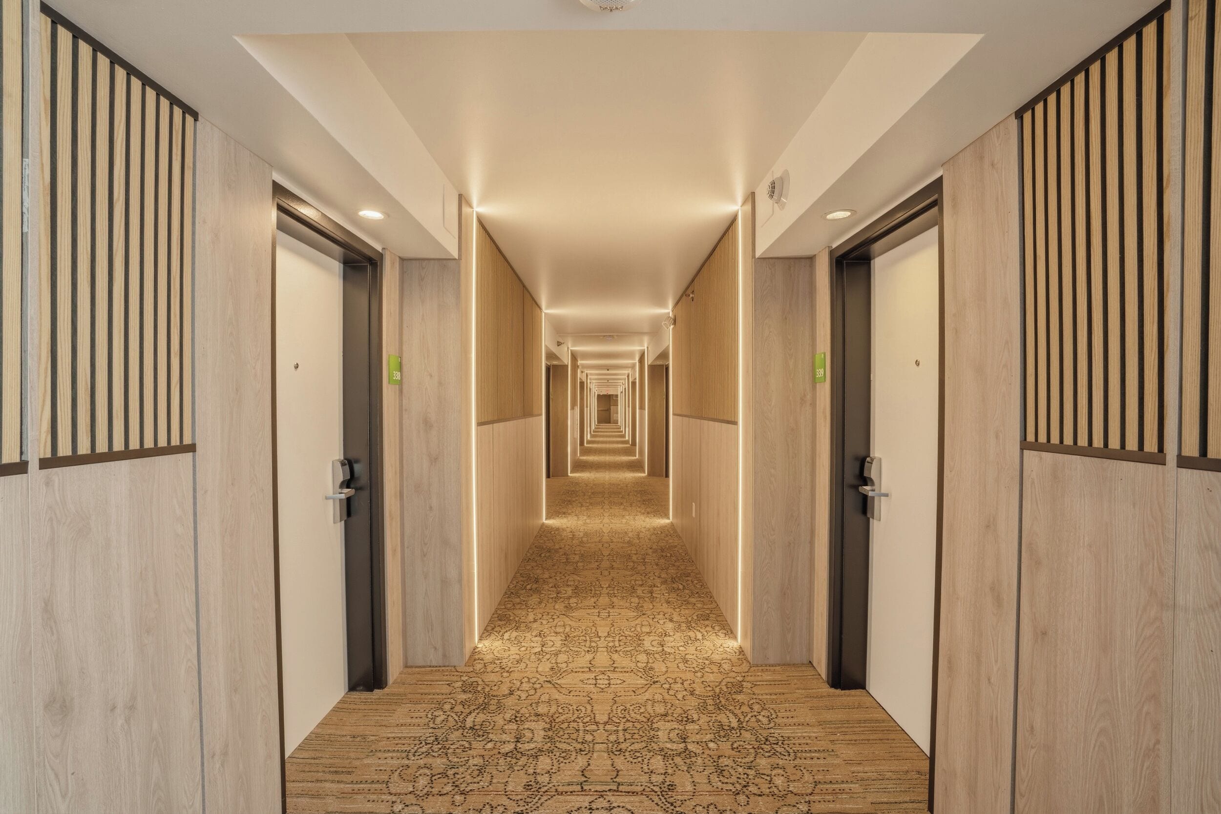 A hotel corridor with wooden paneling, a patterned carpet, and doors marked with room numbers.