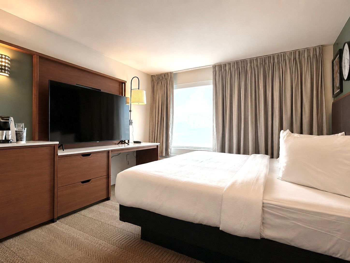 A hotel room with a large bed, a flat-screen TV, a desk with a coffee maker, and a window with curtains.