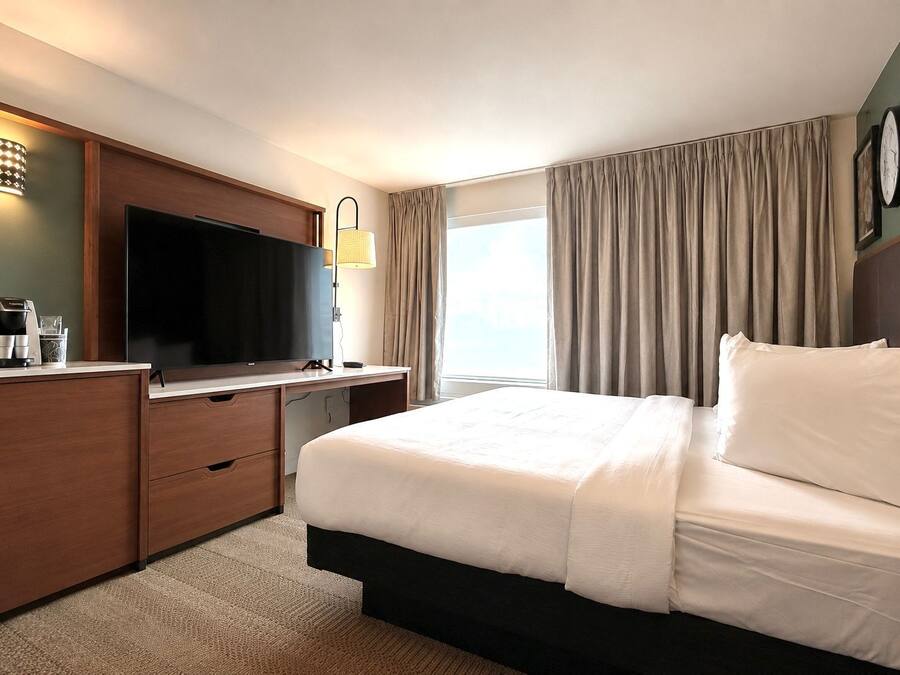 Habitación de hotel con una cama grande, un televisor de pantalla plana, un escritorio con cafetera y una ventana con cortinas.