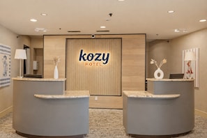 Zona de recepción de un hotel con dos mostradores curvos, un panel de madera con el nombre del hotel y una alfombra estampada.