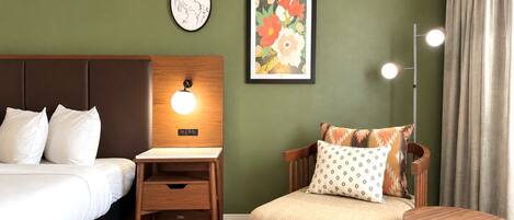 Una habitación de hotel con una cama, un escritorio con lámpara, una silla y una pared decorada con obras de arte.