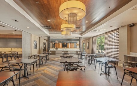 A spacious dining area with multiple tables and chairs, a kitchen in the background, and large windows with curtains.