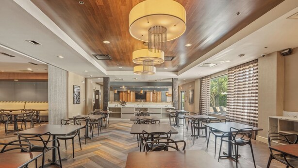 A spacious dining area with multiple tables and chairs, a kitchen in the background, and large windows with curtains. - Kozy Hotel Dallas DFW Airport Area West (Bedford)