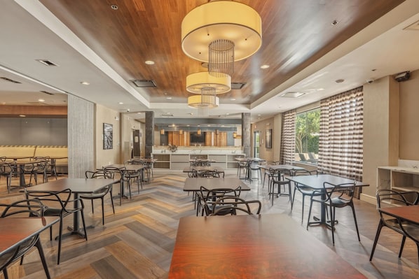A spacious dining area with multiple tables and chairs, a kitchen in the background, and large windows with curtains.