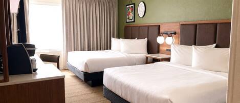 A hotel room with two beds, a desk with a chair, a coffee machine, a clock on the wall, and a window with curtains.