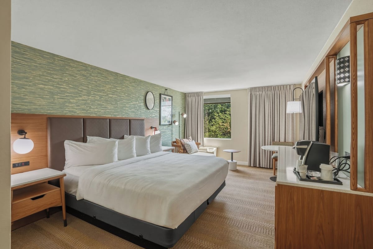A hotel room with a large bed, a desk, a chair, a small table, a lamp, and a window with curtains.