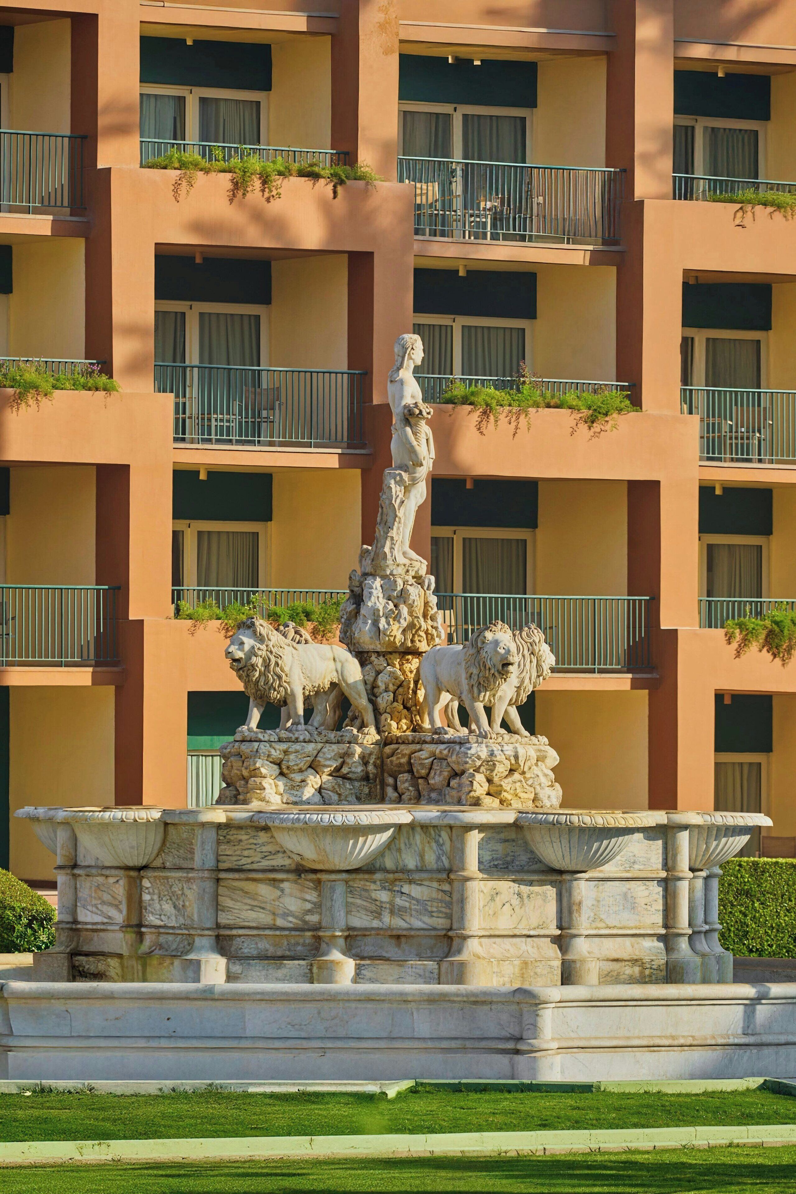A marble fountain with lions and a statue in front of a multi-story building with balconies.