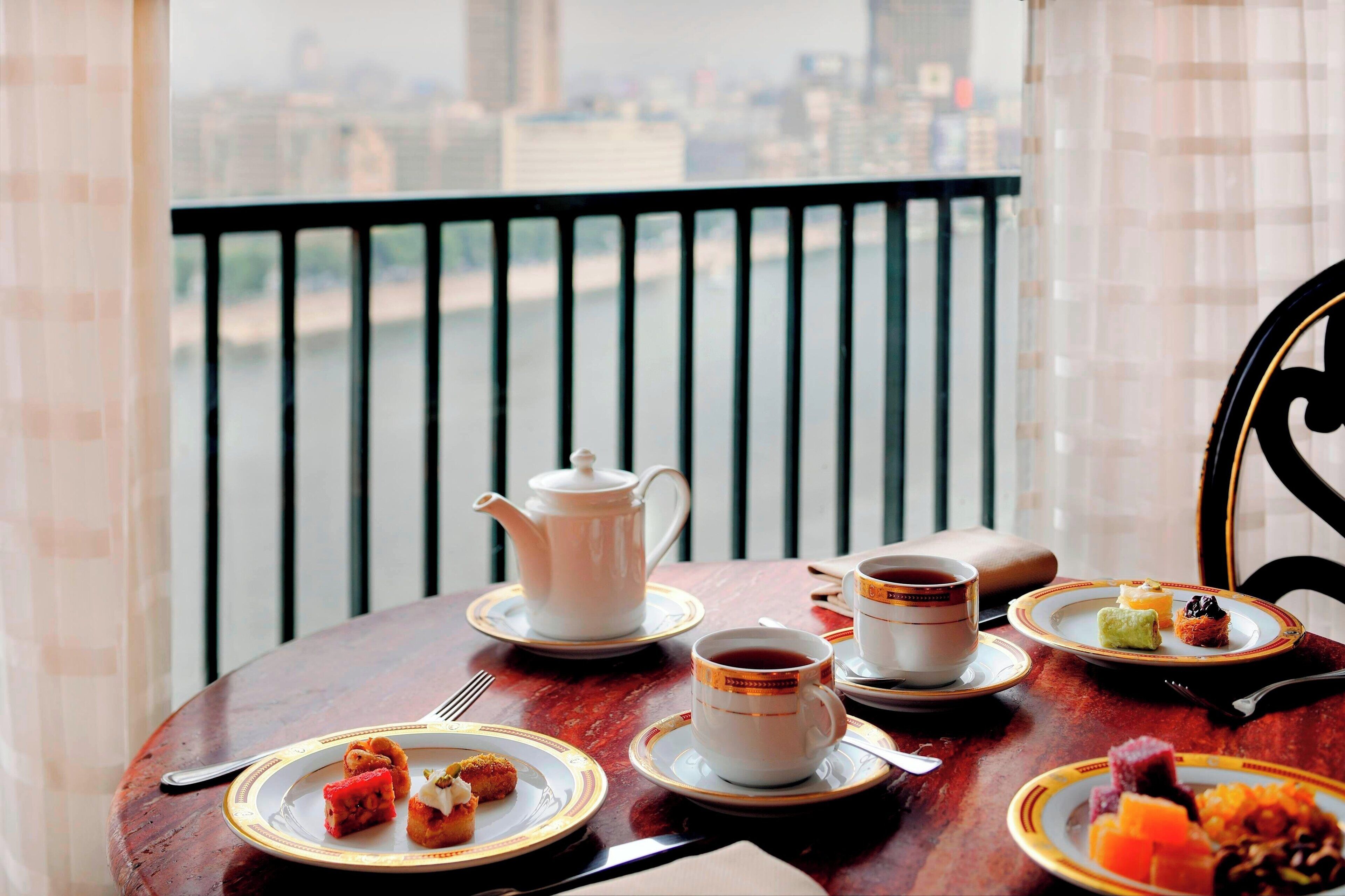 Une table à manger, dressée sur un balcon offrant une vue sur la ville, est garnie d’assiettes remplies de nourriture, d’une théière et de tasses.