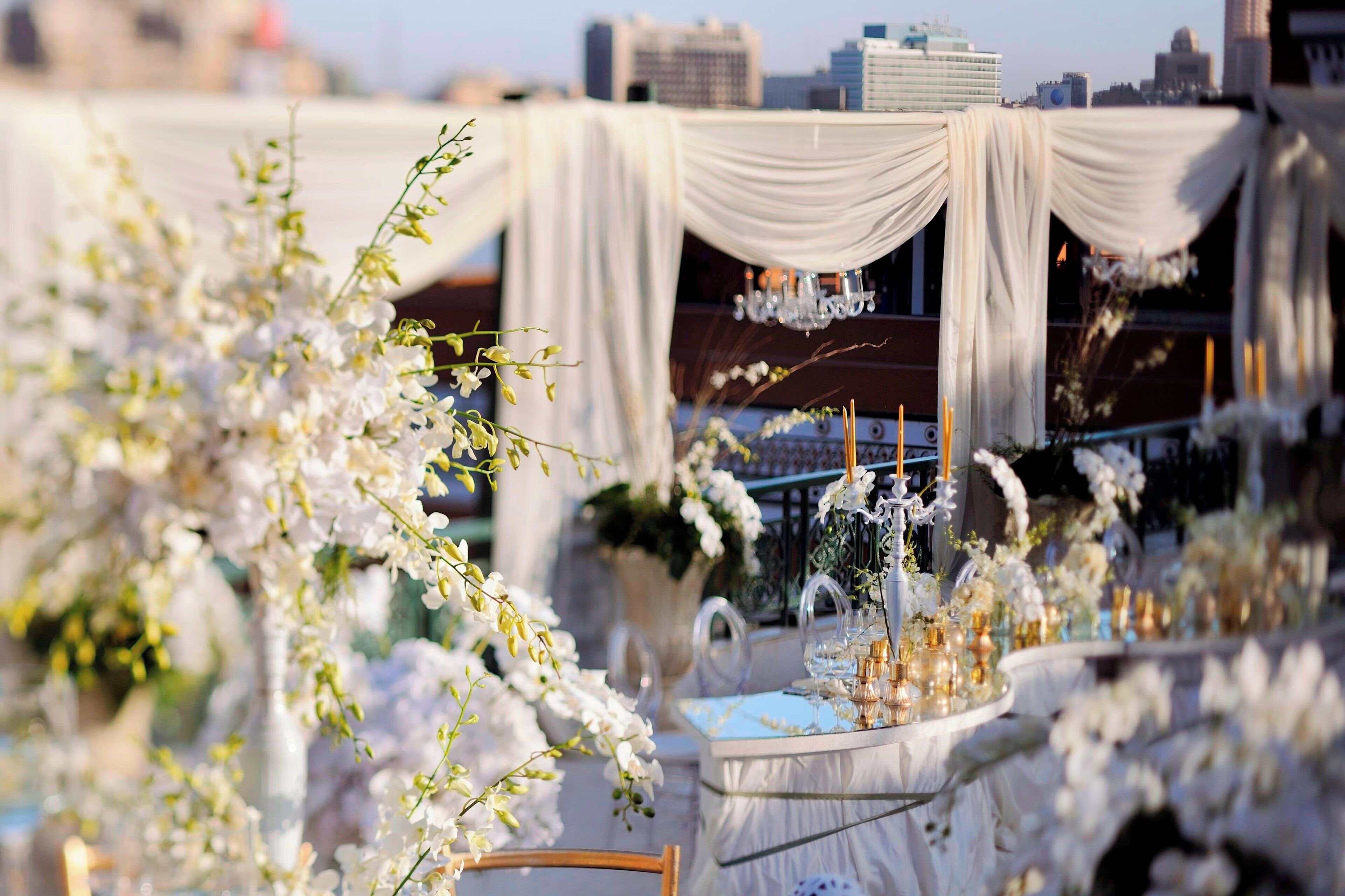 A table set with white flowers and candles, draped with white fabric, in an outdoor setting with a cityscape in the background.