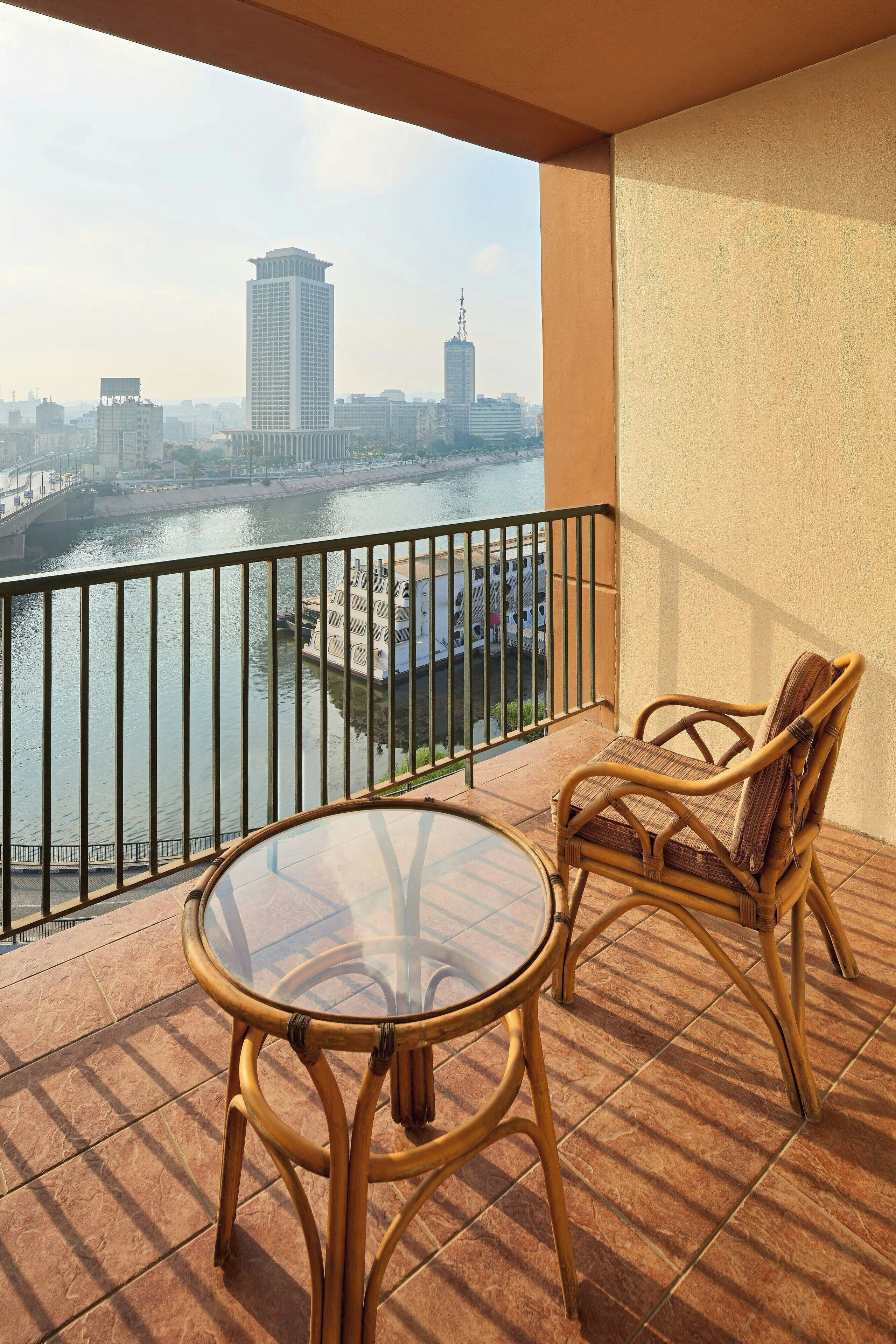 A balcony with a glass table and wicker chairs, overlooking a river with buildings in the background.