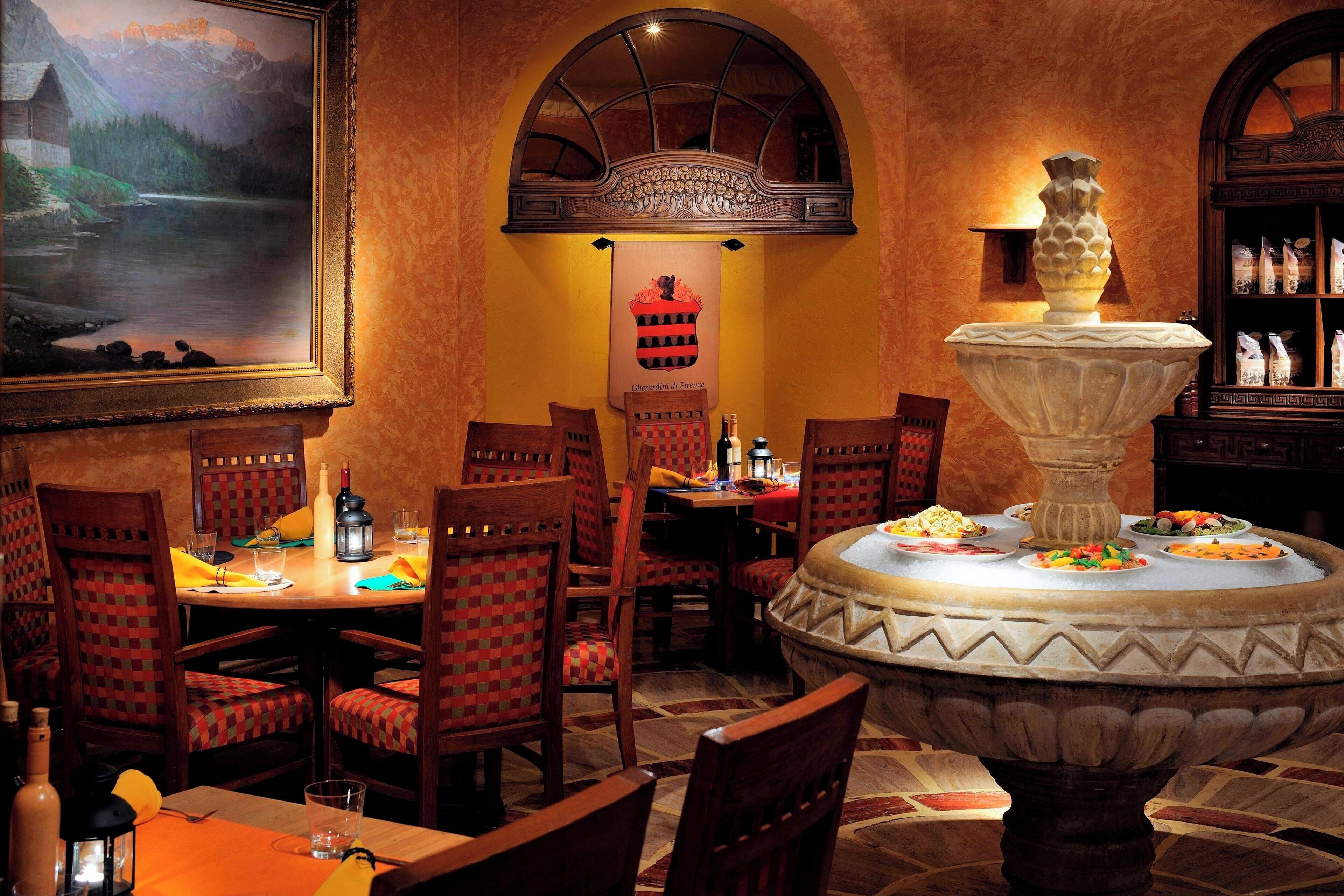 A dining area with wooden chairs, a table with a bottle and glasses, a painting on the wall, and a fountain centerpiece.