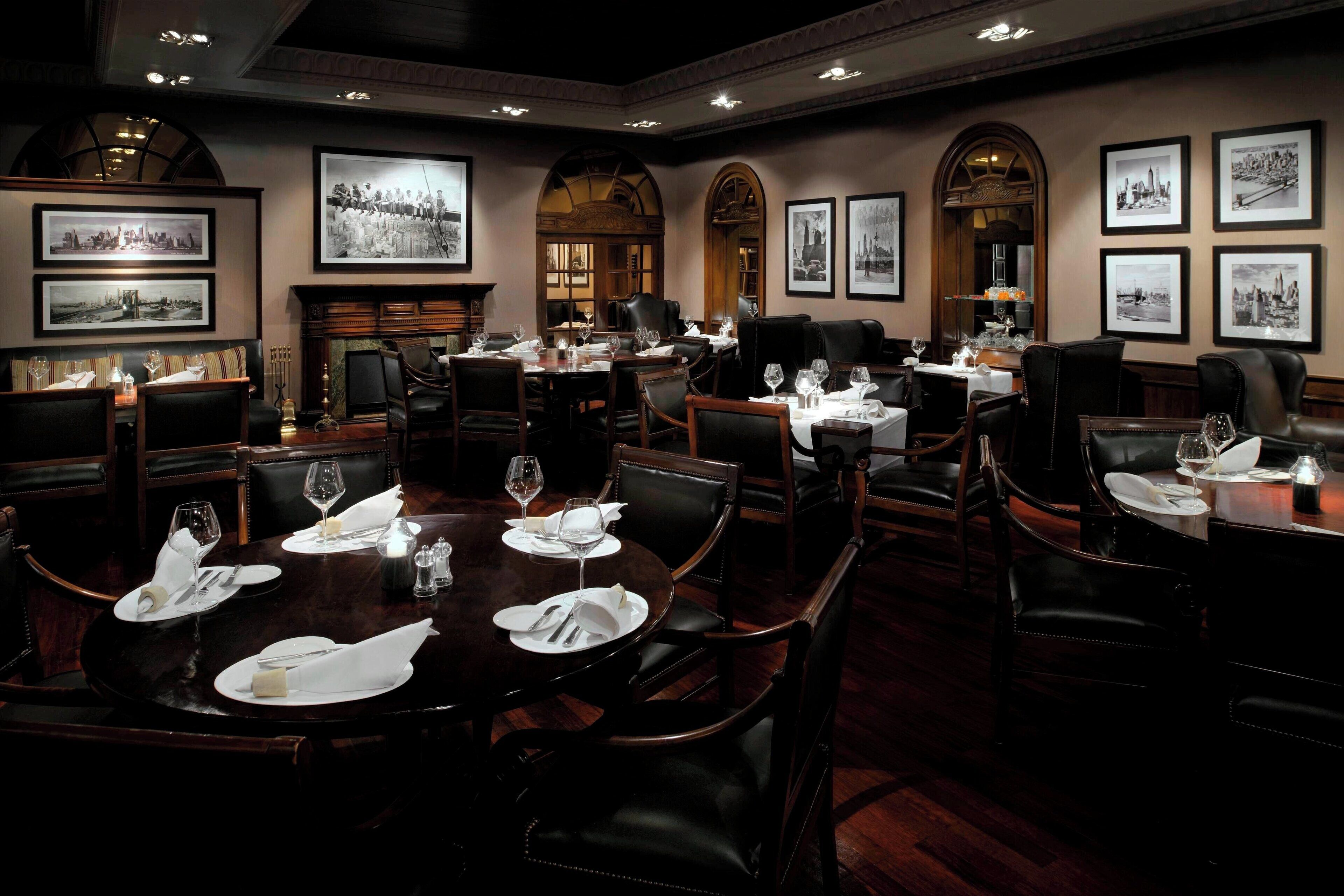 A formal dining room with multiple tables set for service, framed historical photographs on the walls, and a classic fireplace.