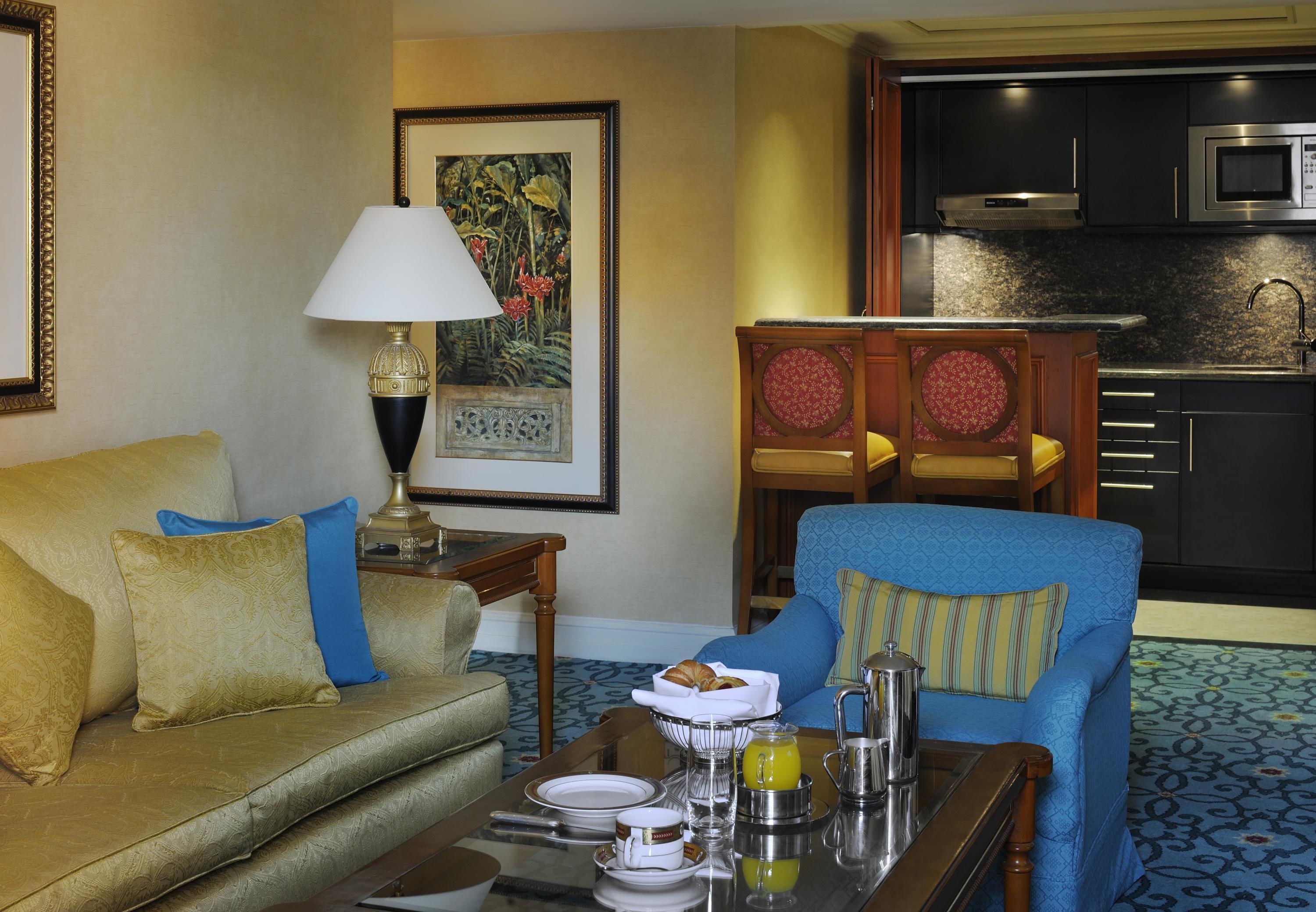 A hotel room with a sofa, armchair, dining table with breakfast items, a lamp, a painting, and a kitchen area.