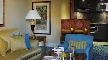 Habitación de hotel con sofá, sillón, mesa de comedor con alimentos para el desayuno, una lámpara, un cuadro y una zona de cocina.
