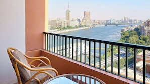 A balcony with wicker chairs and a glass table, overlooking a river with buildings and a bridge in the background.