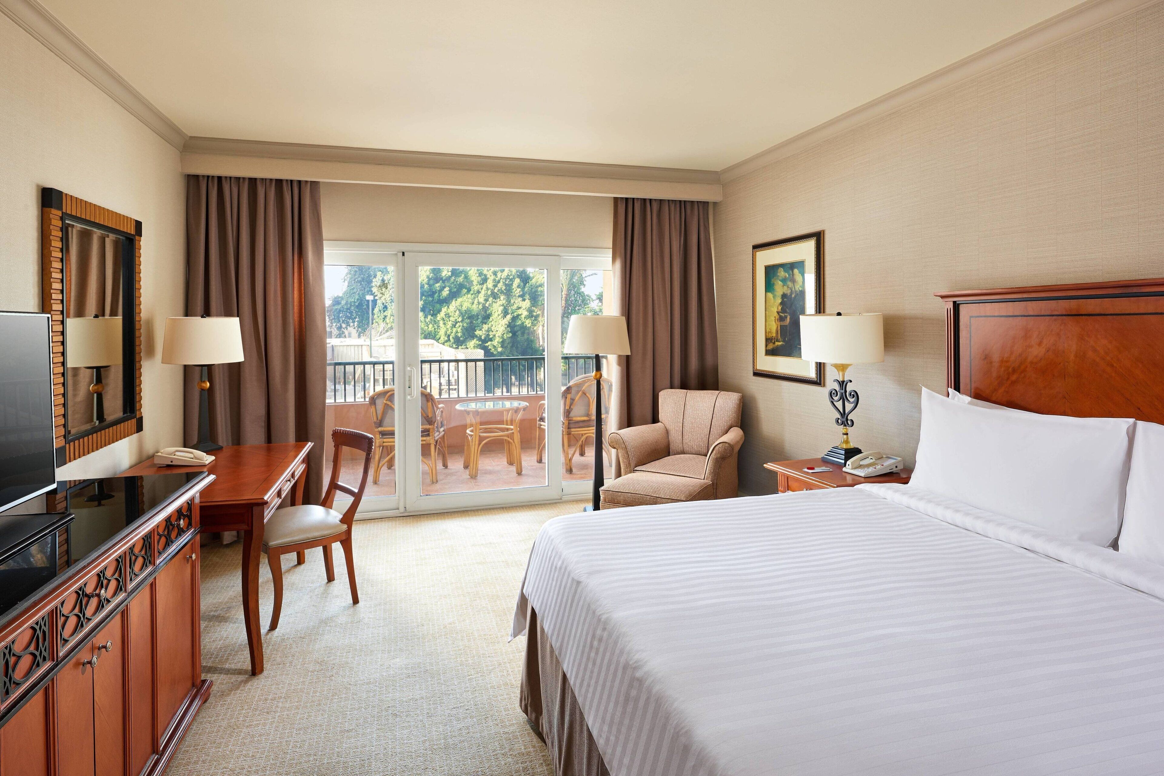 A hotel room with a bed, a desk with a chair, a television, a telephone, and a balcony with outdoor furniture.