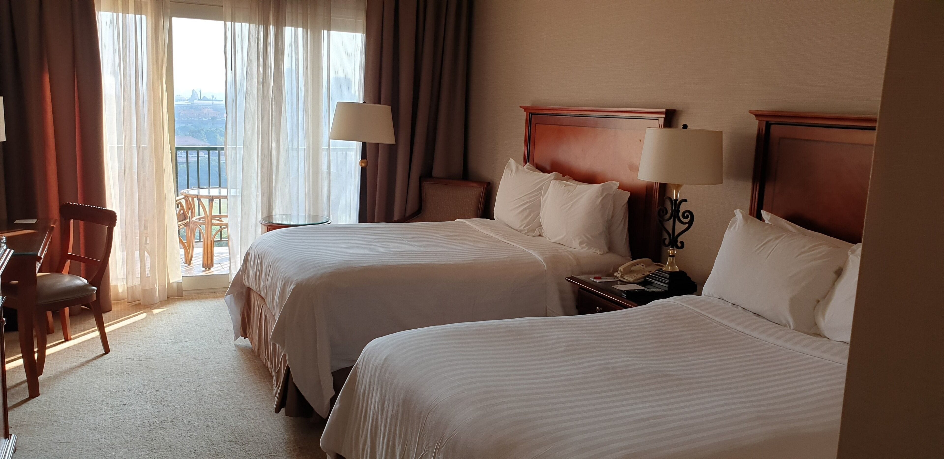 A hotel room with two beds, a chair, a table, and a balcony with curtains.