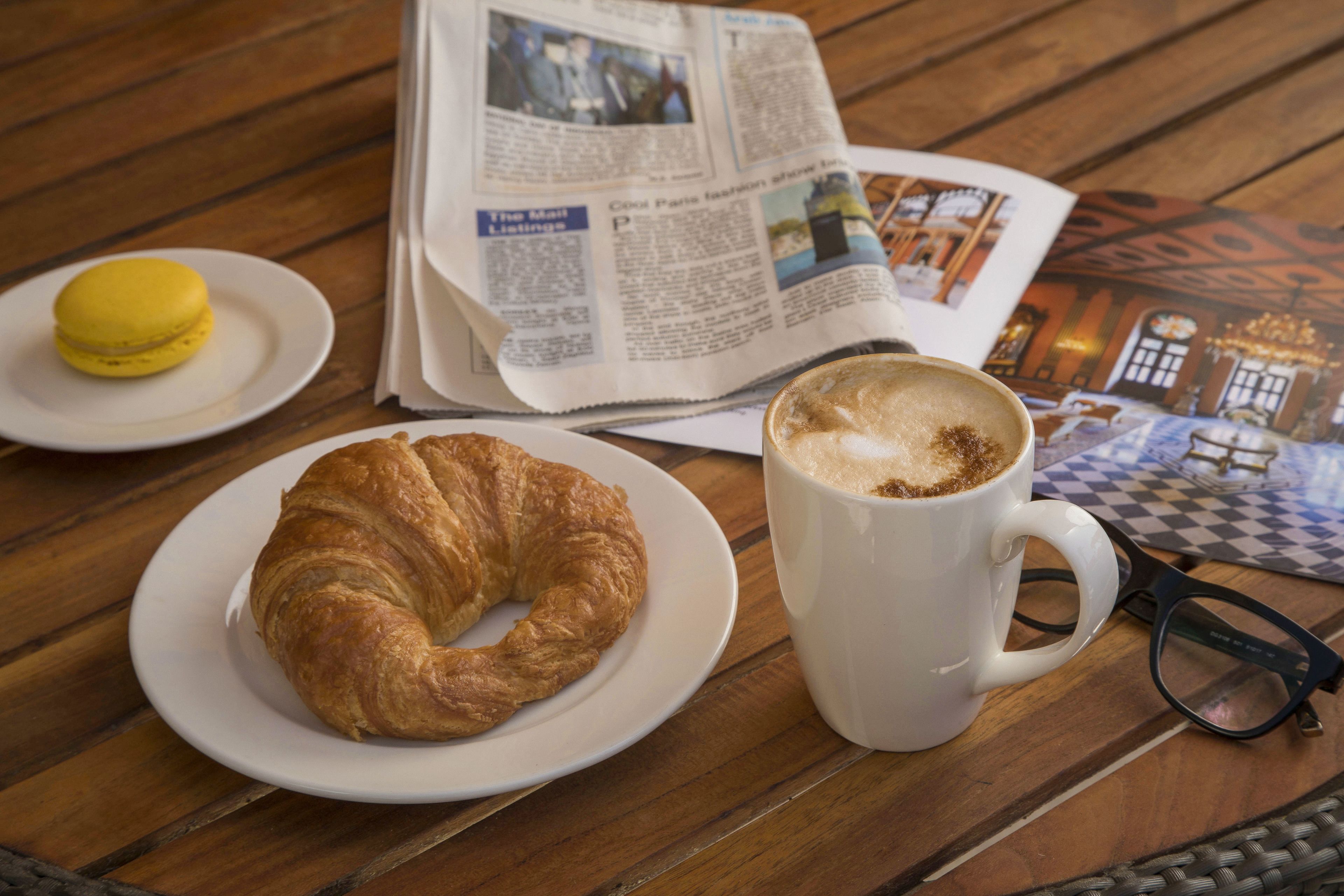 Un croissant su un piatto, una tazza di caffè, un paio di occhiali e un giornale su un tavolo di legno.