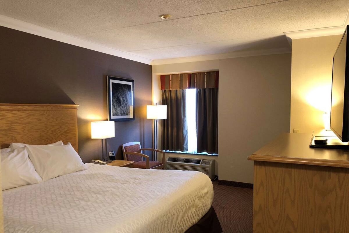 A hotel room with a large bed, two bedside lamps, a chair, a desk with a lamp, a window with curtains, and a framed picture on the wall.