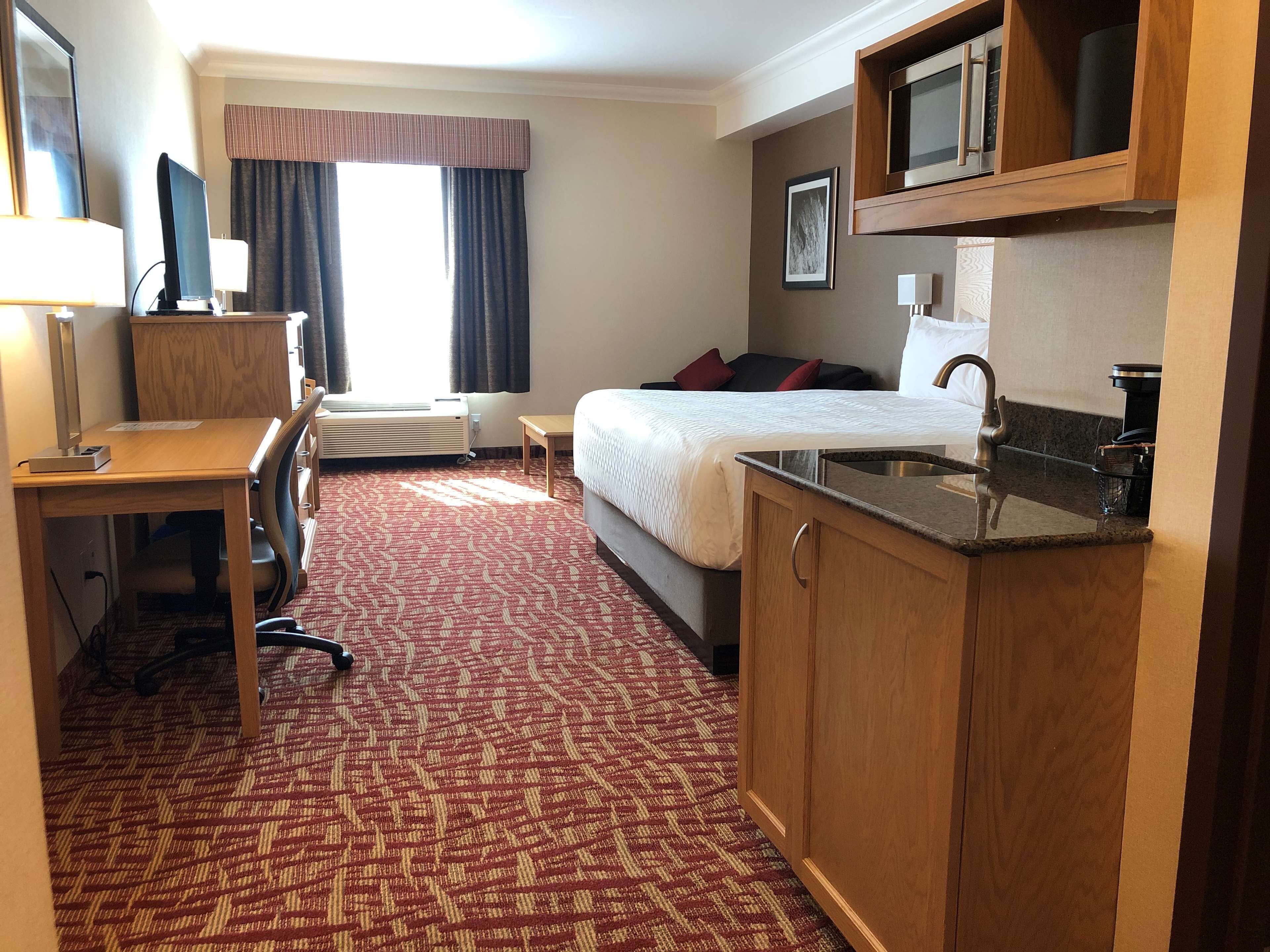 A hotel room with a desk, a bed, a kitchenette, and a large window with curtains.