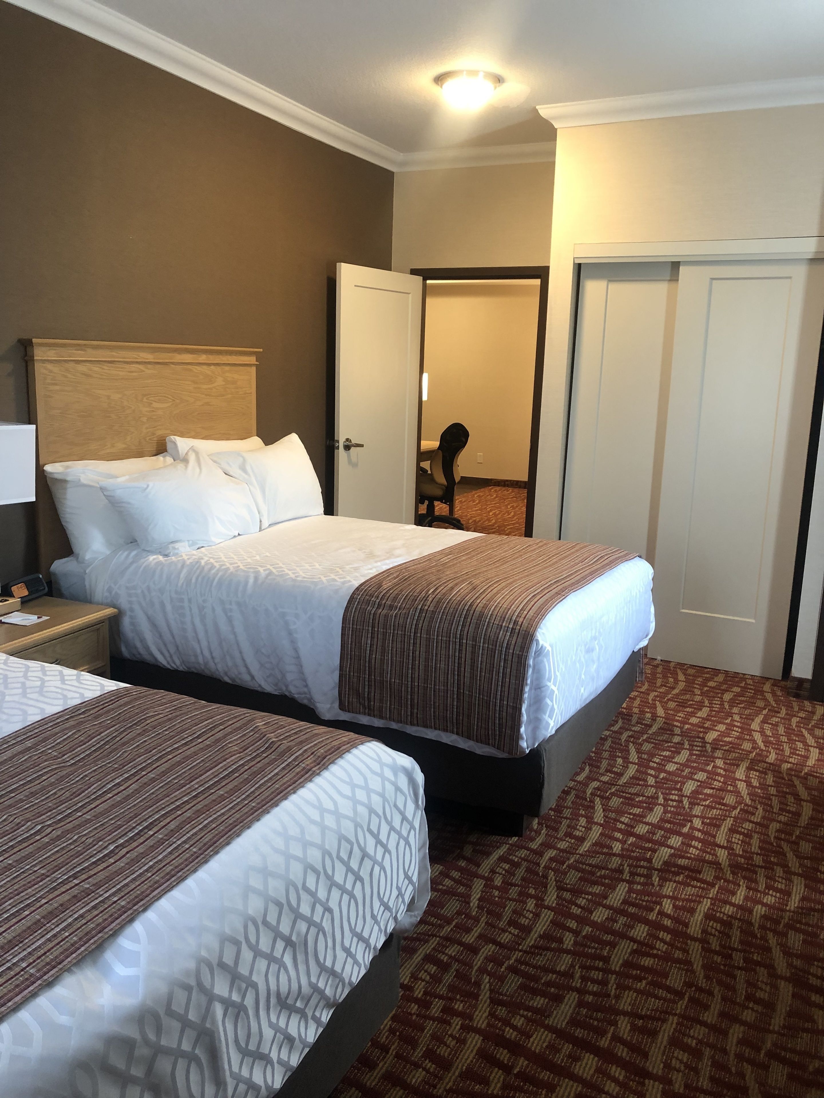 Una habitación de hotel con dos camas, una mesita de noche, una lámpara y una puerta que da a otra habitación.