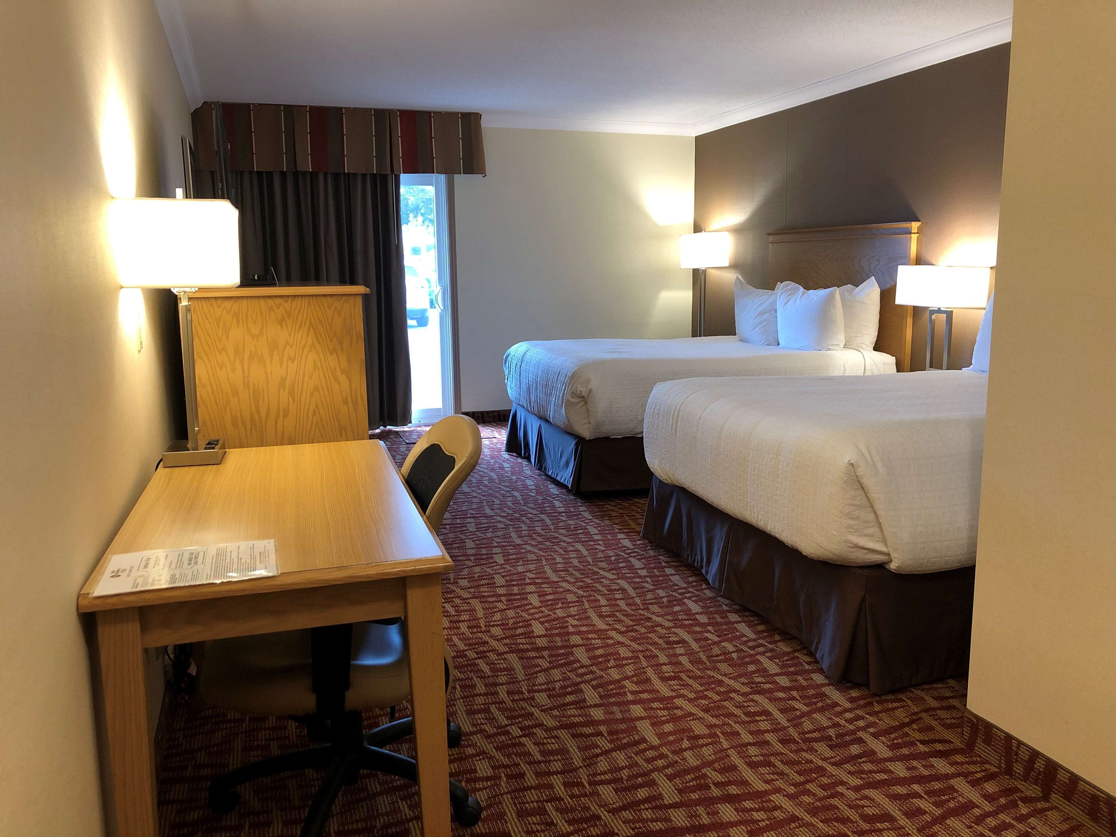 Une chambre d’hôtel avec deux lits, un bureau et une chaise. Il y a des lampes de chaque côté des lits et une fenêtre avec des rideaux.