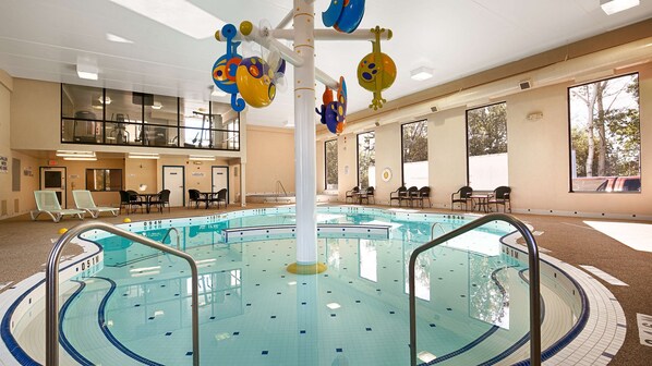 An indoor swimming pool with a central pole, lounge chairs, and a view of the outdoors through large windows.