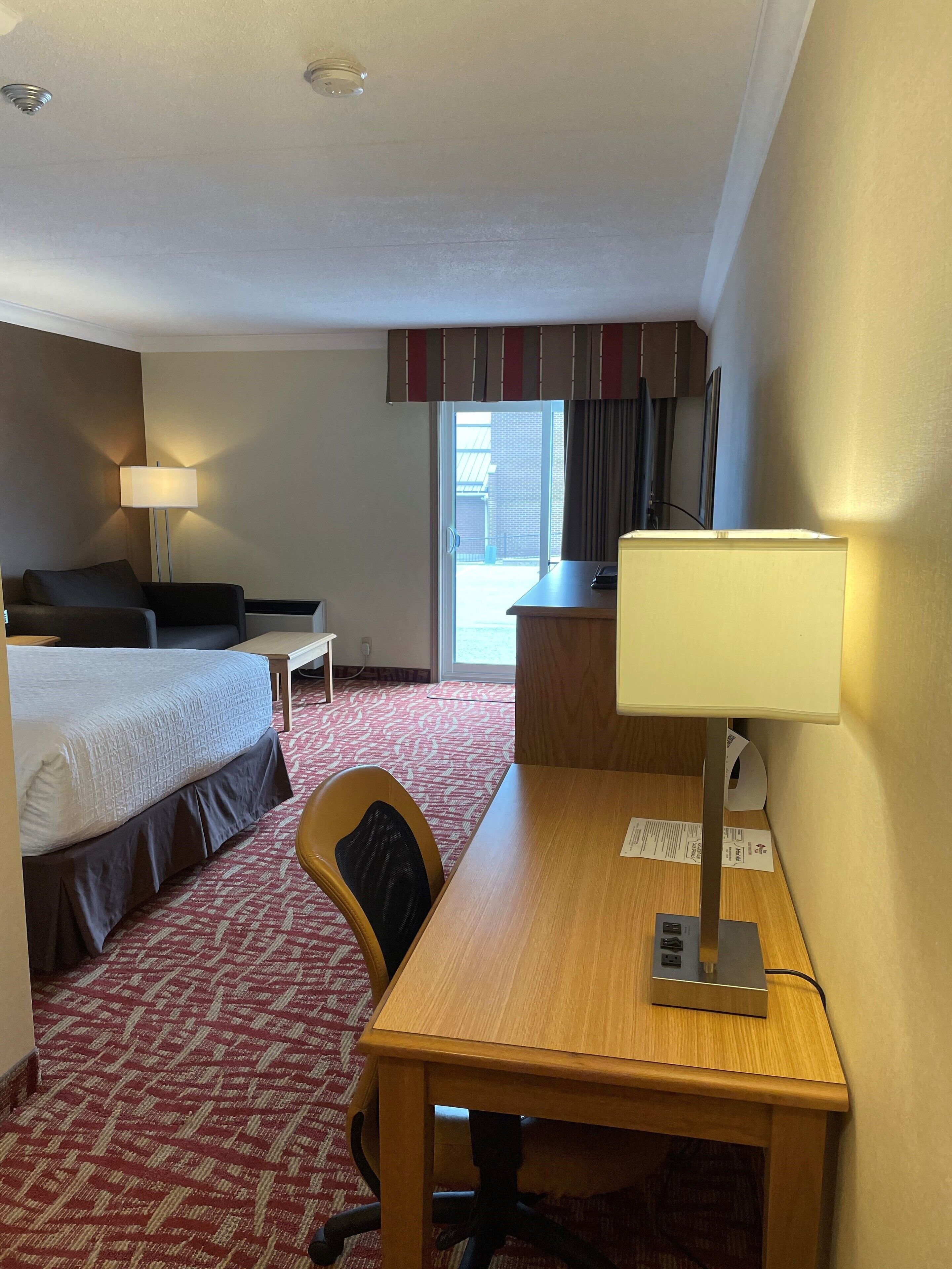 A hotel room with a bed, a desk with a lamp, a chair, and a view of the outside.