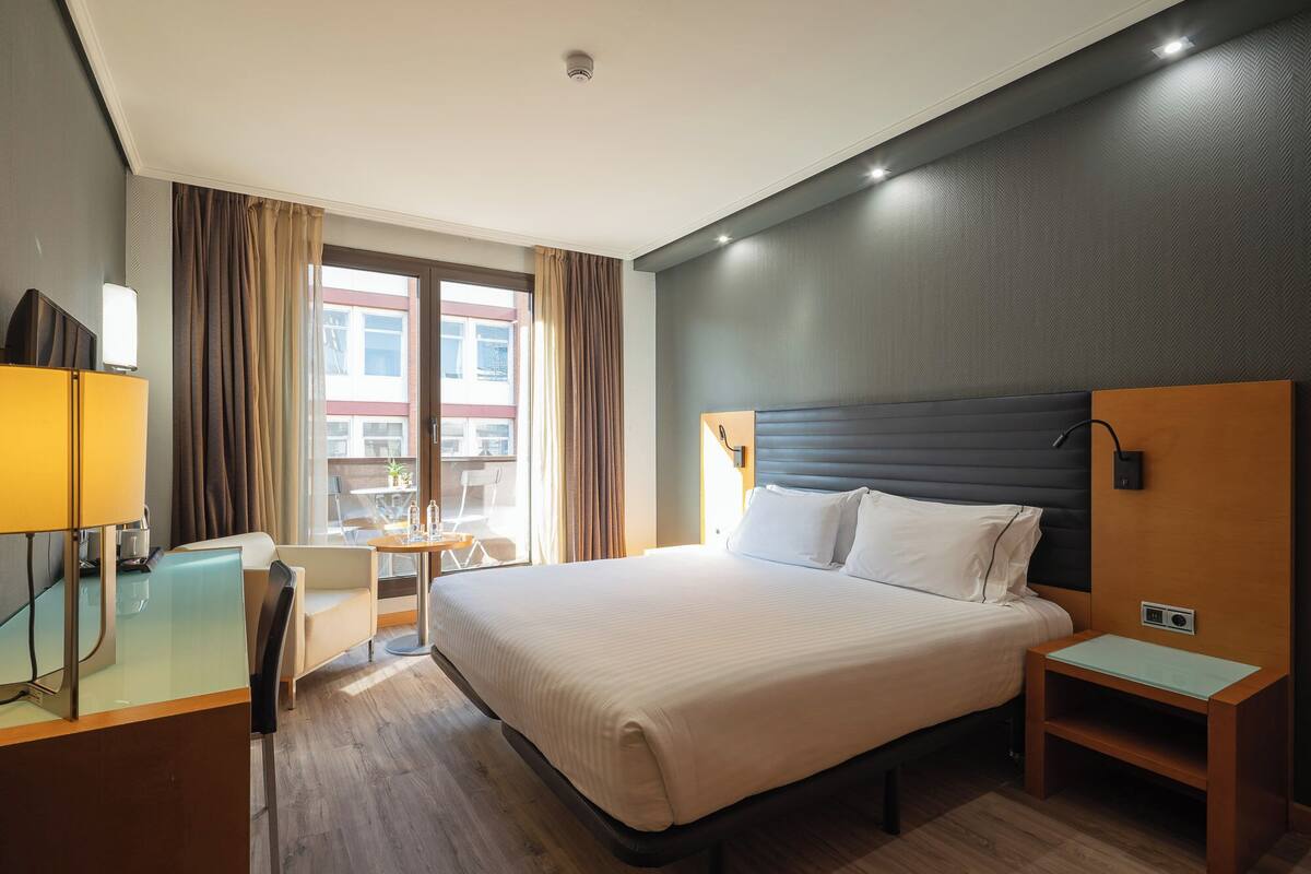 Hotellihuone, jossa on suuri sänky, työpöytä lampun kanssa, pieni pöytä tuoleineen ja ikkunasta avautuva näkymä rakennuksiin.