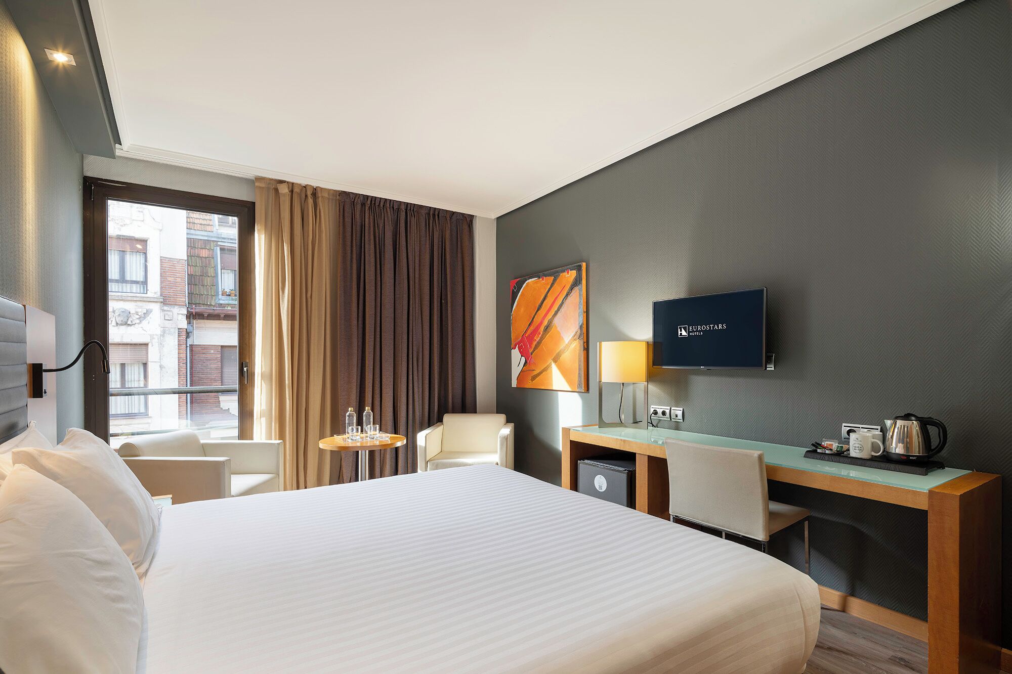 A hotel room with a bed, a desk, a chair, a TV, and a window with curtains.