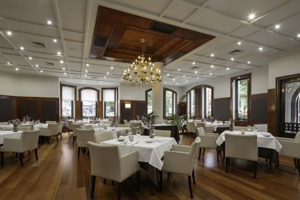 A spacious dining area with multiple tables and chairs, a chandelier, and large windows allowing natural light.