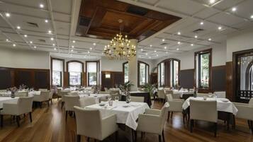 A spacious dining area with multiple tables and chairs, a chandelier, and large windows allowing natural light.