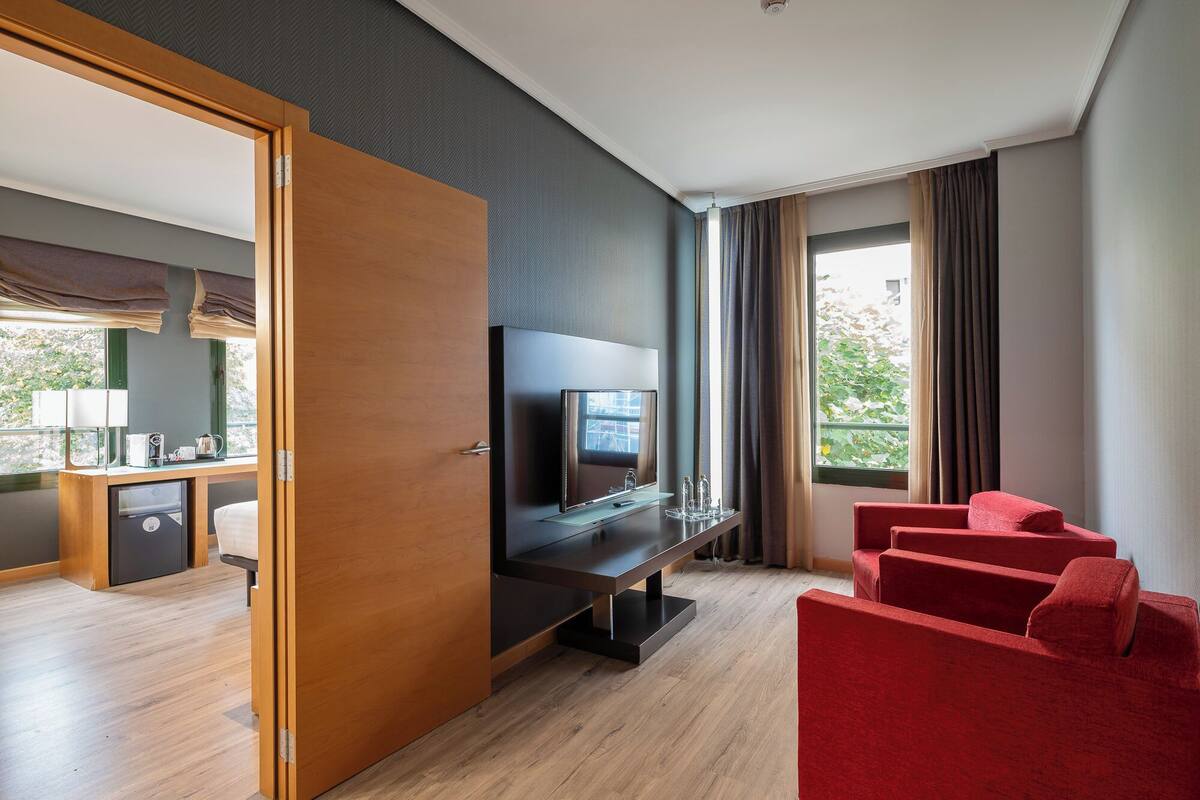 Moderni hotellihuone, jossa on puulattia, taulutelevisio, punainen sohva sekä ruokailutila, jossa on pöytä ja tuolit.