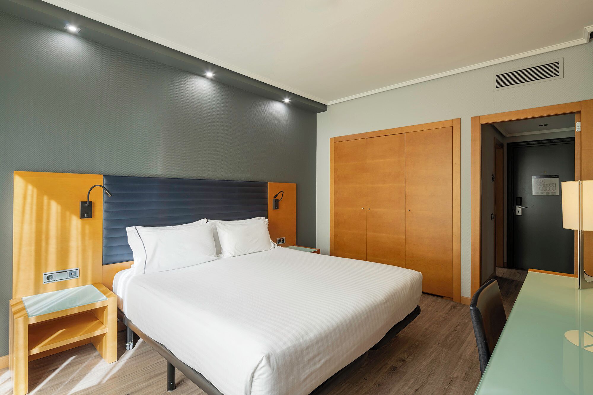 A hotel room with a bed, bedside table, wardrobe, and a desk with a chair.