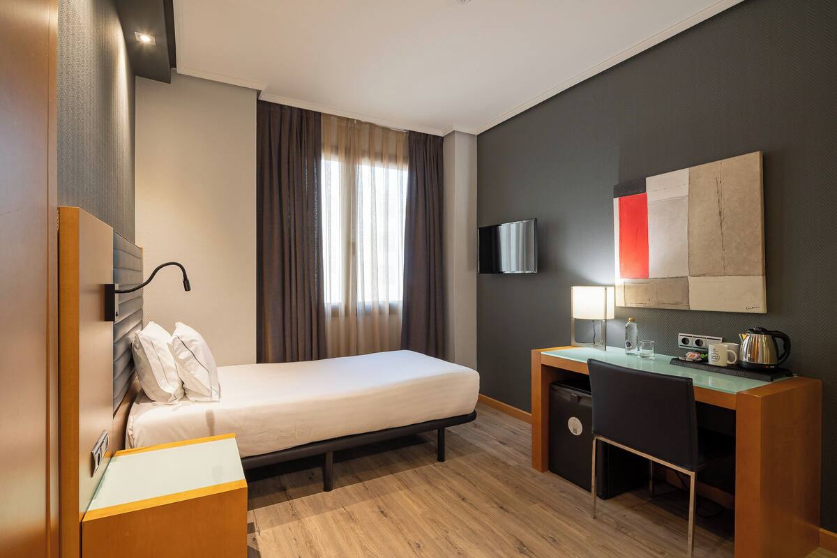 Hotellihuone, jossa on sänky, työpöytä, tuoli, televisio ja suuri ikkuna, jossa on verhot.