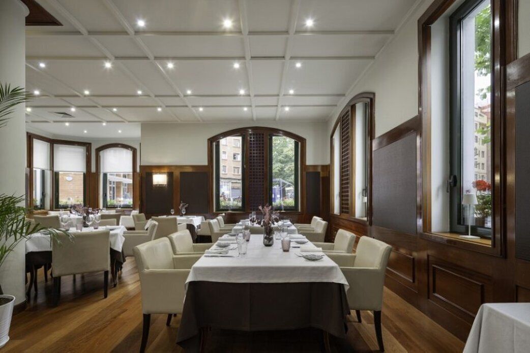 Un comedor espacioso con varias mesas dispuestas para comer, que cuenta con un techo alto con iluminación empotrada y grandes ventanas que ofrecen luz natural.