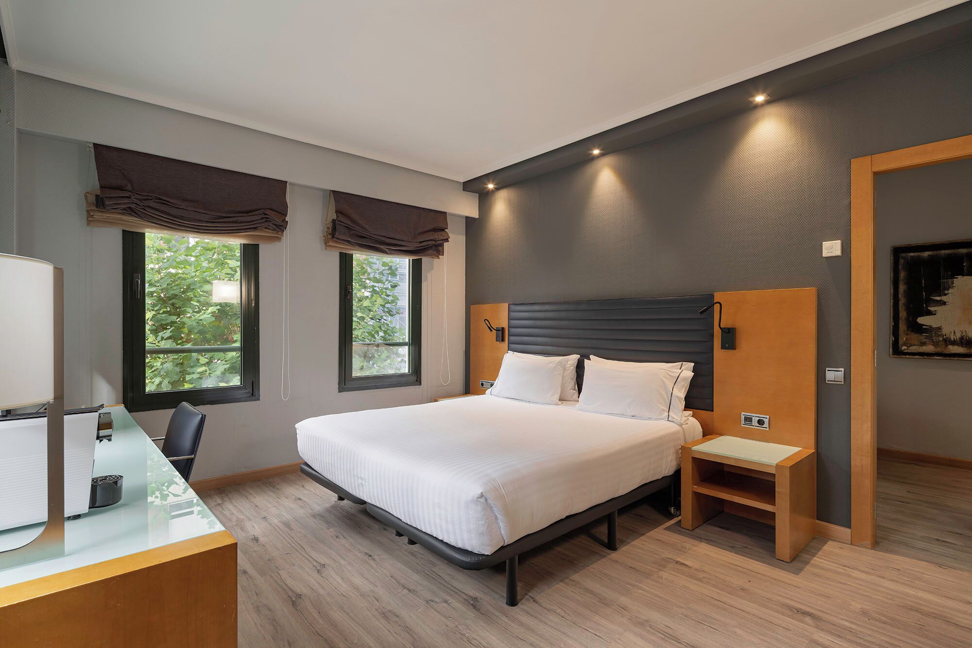 Una habitación de hotel moderna con una cama grande, mesitas de noche, un escritorio con lámpara y ventanas con cortinas.