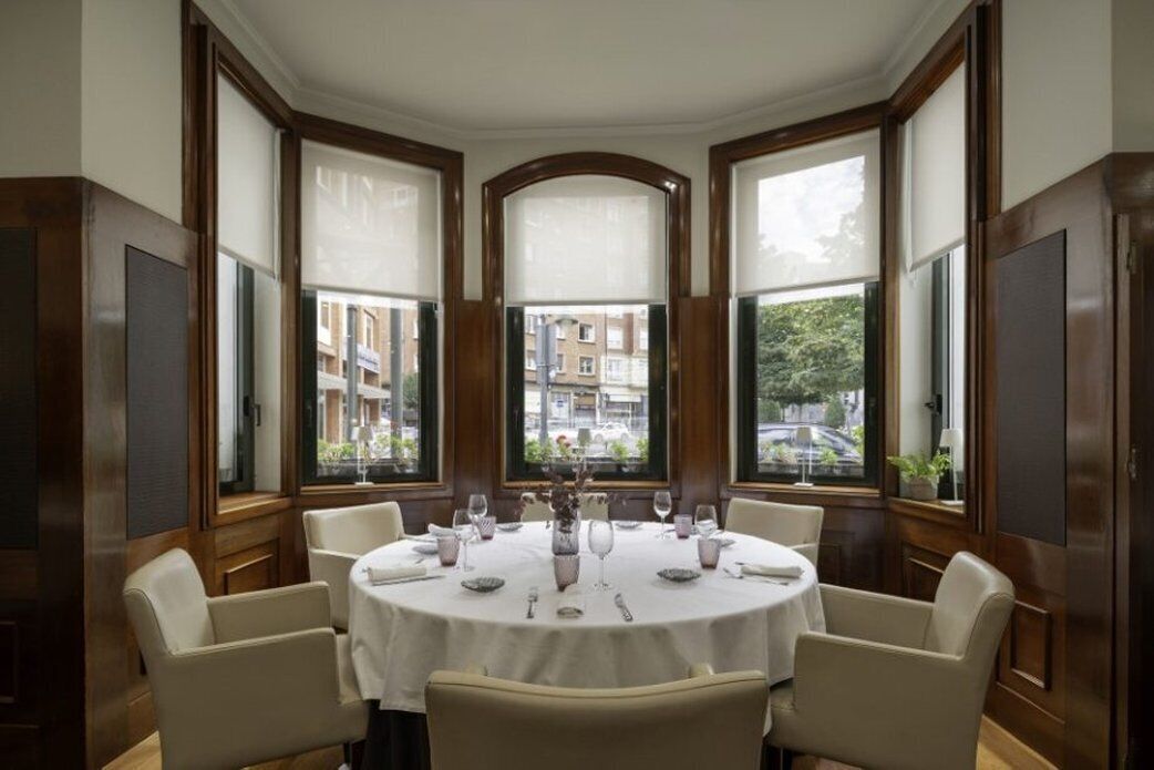 Una mesa redonda para comer con seis sillas, dispuesta para una comida, en una habitación con ventanas grandes y paneles de madera.