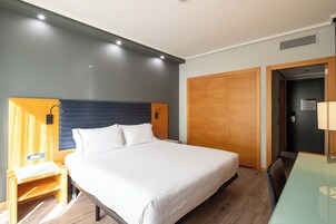 Ein Hotelzimmer mit Bett, Nachttisch, Lampe, Schrank und Schreibtisch mit Stuhl.