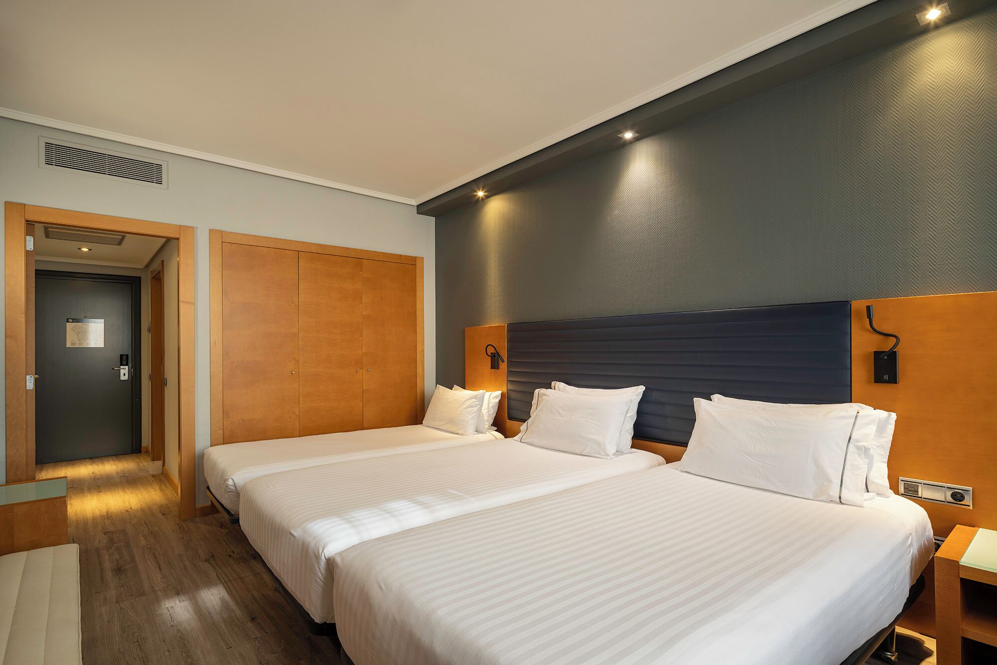 A hotel room with two beds, a wooden wardrobe, a door with a sign, and a wall-mounted light fixture.