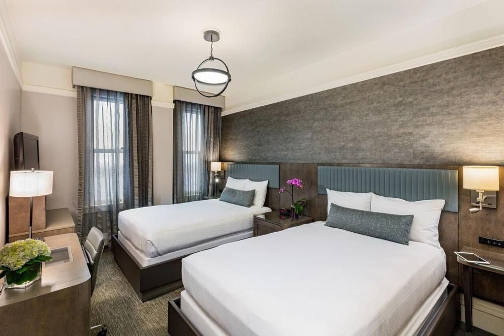 A hotel room with two beds, a desk, a chair, a lamp, and a vase with flowers.