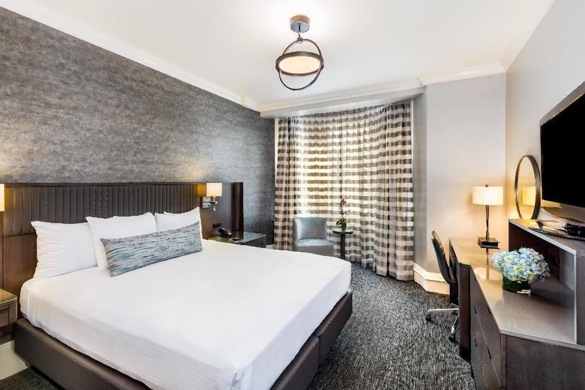 A hotel room with a large bed, a desk, a chair, a television, and a window with curtains.