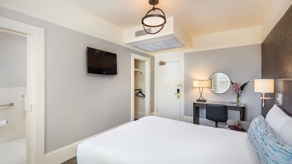 Habitación de hotel con una cama grande, un televisor de pantalla plana en la pared, un escritorio con una silla, un espejo redondo y una lámpara de noche.