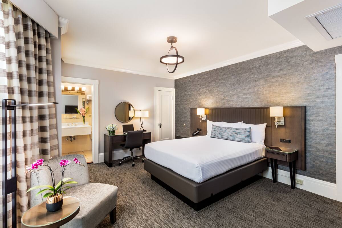 Habitación de hotel con una cama grande, un escritorio con silla, una pequeña mesa con flores, un espejo y un baño visible a través de una puerta abierta.