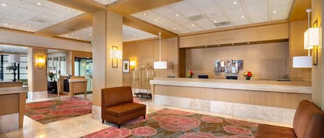 A hotel lobby with a reception desk, two armchairs, a patterned carpet, and a ceiling with recessed lighting.